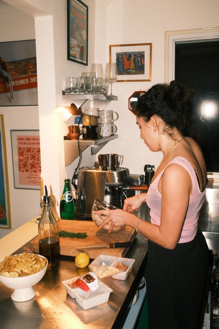 Image of a person in a pink top and black pants cooking in a small kitchen with stainless steel countertops and white walls.