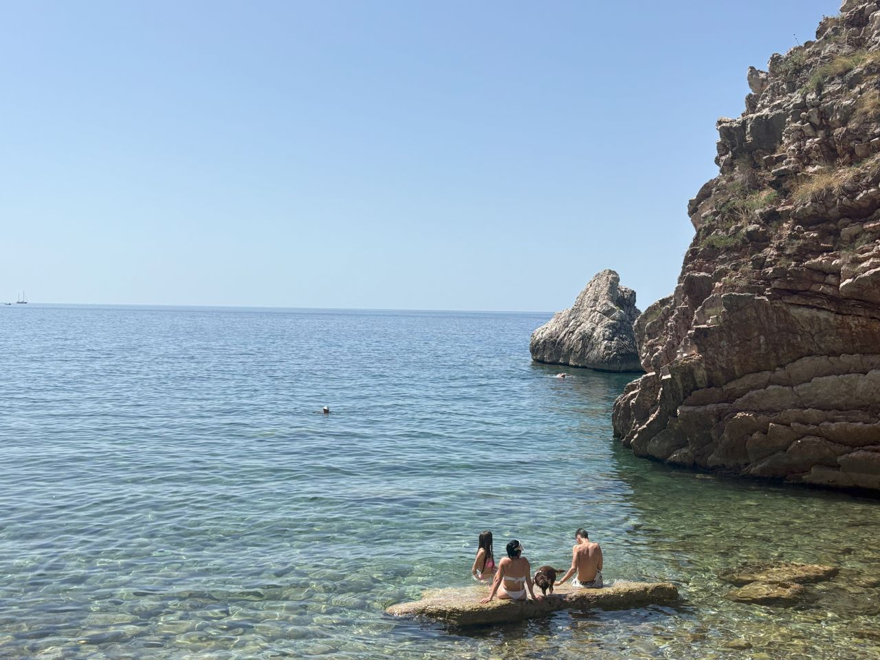 A sun-bathed beach with rocks and people bathing in crystal-clear, blue sea.