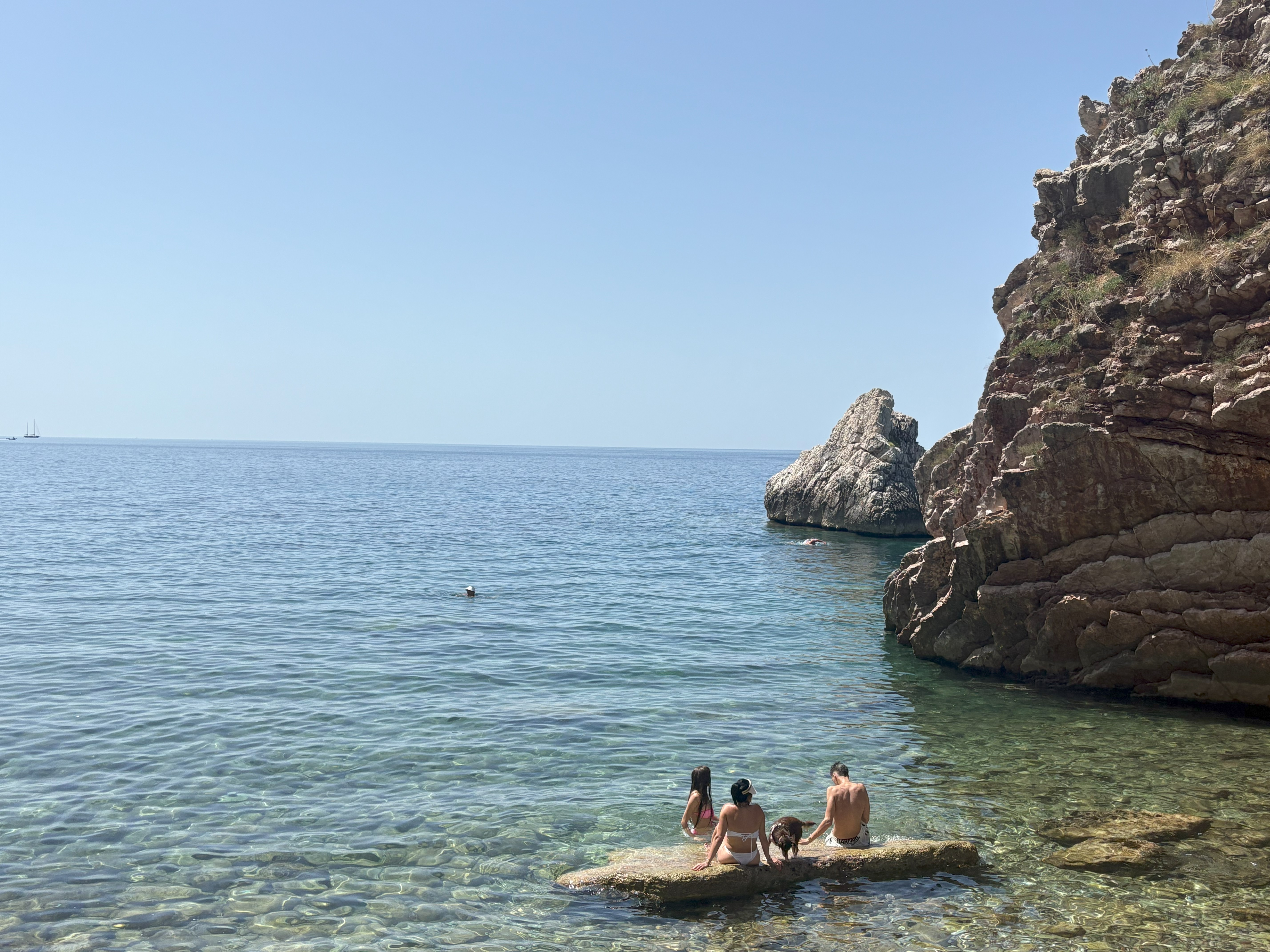 A sun-bathed beach with rocks and people bathing in crystal-clear, blue sea.