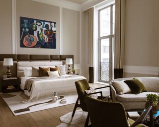 neutral bedroom with bed with oversized headboard, colorful statement artwork and living area with curved sofa and olive chairs