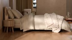 A modern, minimalist bedroom with a large made double bed with cream bedding. The floor is wood, and to the right of the bed, in the foreground, is a circular bedside table with two vases of dried flowers. In the background is a large window with a drawn brown curtain.