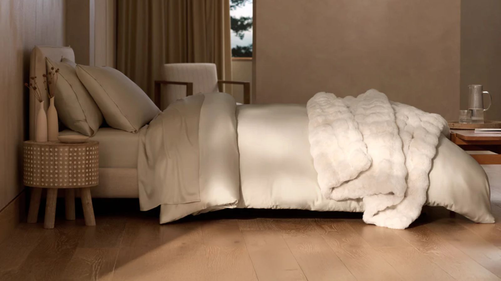 A modern, minimalist bedroom with a large made double bed with cream bedding. The floor is wood, and to the right of the bed, in the foreground, is a circular bedside table with two vases of dried flowers. In the background is a large window with a drawn brown curtain.