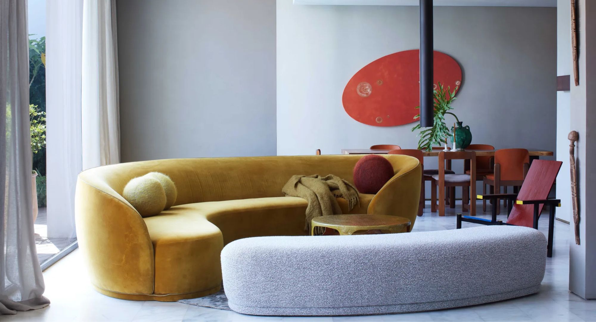 Mustard curved yellow sofa with round cushions, grey curved footstool, red artwork, drapes and wooden table with red chairs