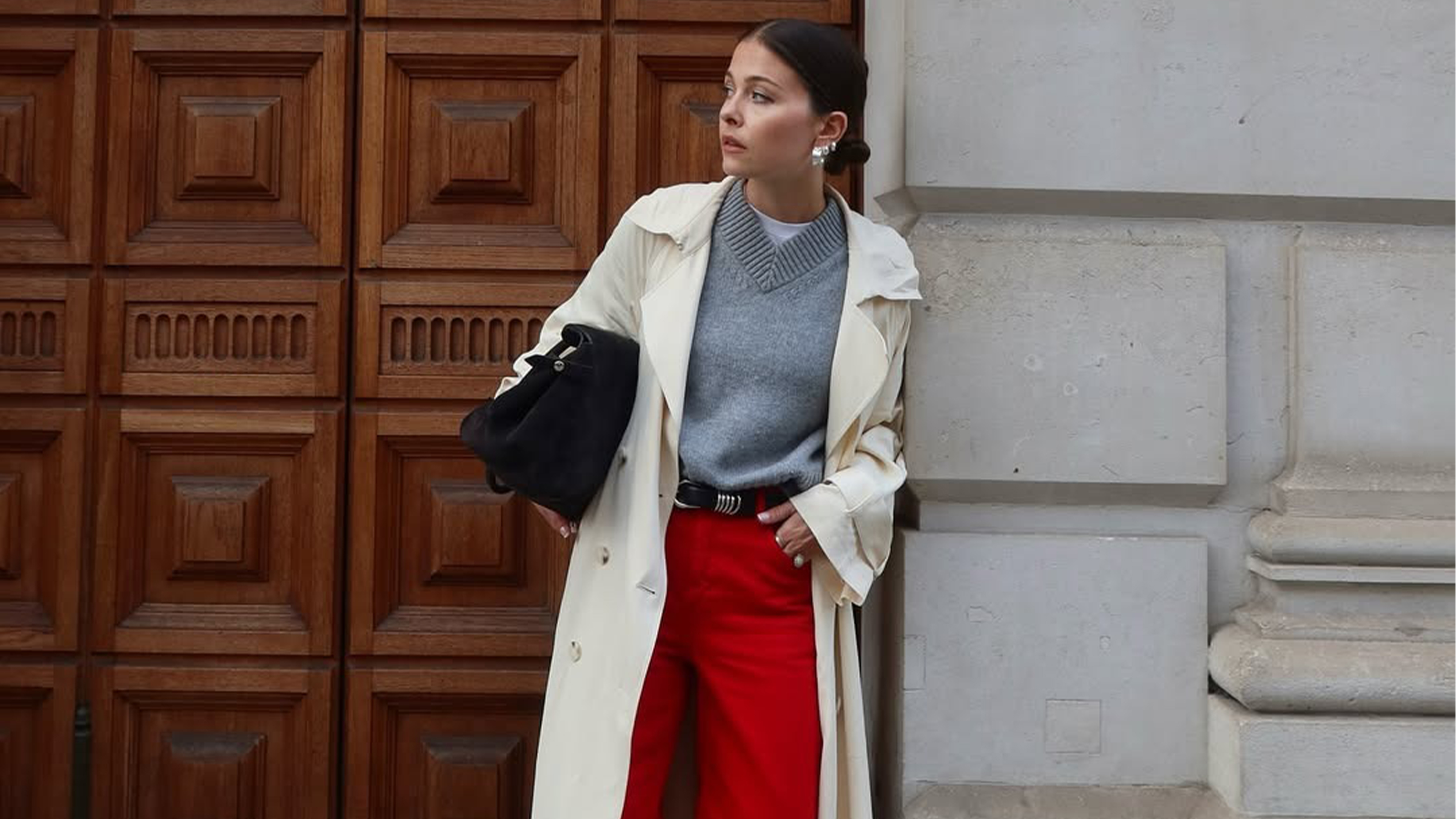 Influencer Alice Satterthwaite wears red jeans, a grey high-V-neck sweater, a long trench coat, pointed-toe boots, and a black suede tote. 