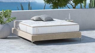 The Saatva Zenhaven Mattress photographed on a beige bedframe on a sunny patio overlooking the ocean