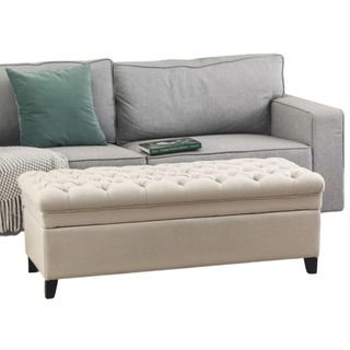 Cream tufted storage ottoman with a grey sofa with green cushions, blanket and book