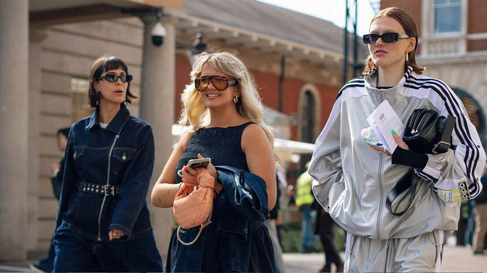 A group of three. women walking. One wears a denim jumpsuit. Another wears a denim dress and adidas. The last one wears a track jacket, shorts, and long boots. 