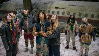 Nat, Van, Taissa, Akilah, Misty, Shauna, Melissa, Travis, and Lottie all bundled up in coats and long pants standing outside the plane fuselage looking up into the sky in Yellowjackets Season 4