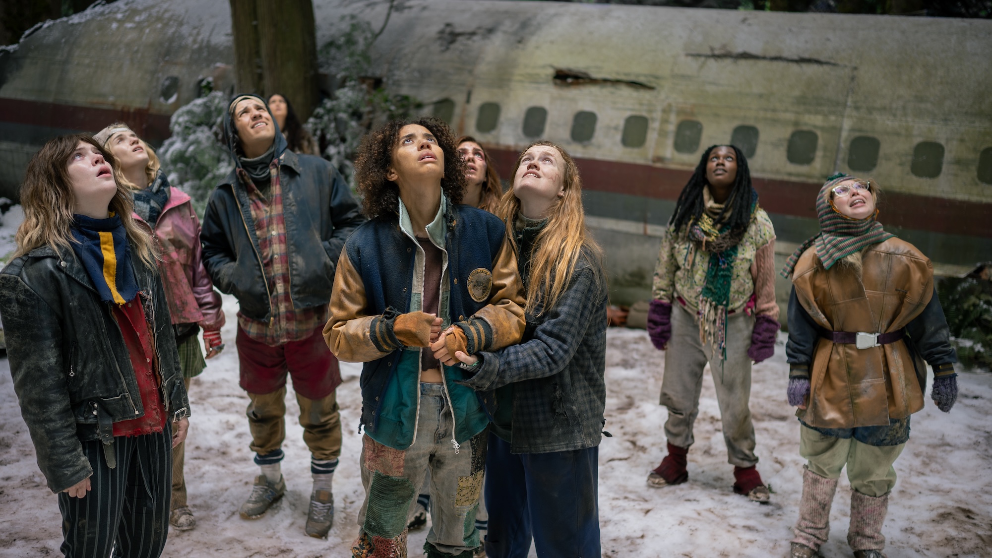 Nat, Van, Taissa, Akilah, Misty, Shauna, Melissa, Travis, and Lottie all bundled up in coats and long pants standing outside the plane fuselage looking up into the sky in Yellowjackets Season 4