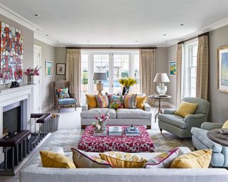 Large sitting room with two gray sofas and two gray armchairs, finished with yellow and pink patterned cushions, surrounding a pink, floral patterned ottoman, gray-beige painted walls with dark yellow and gray checked curtains, traditional fireplace with metal and leather fire front, large pale yellow and blue rug, dark wood, rounded side tables