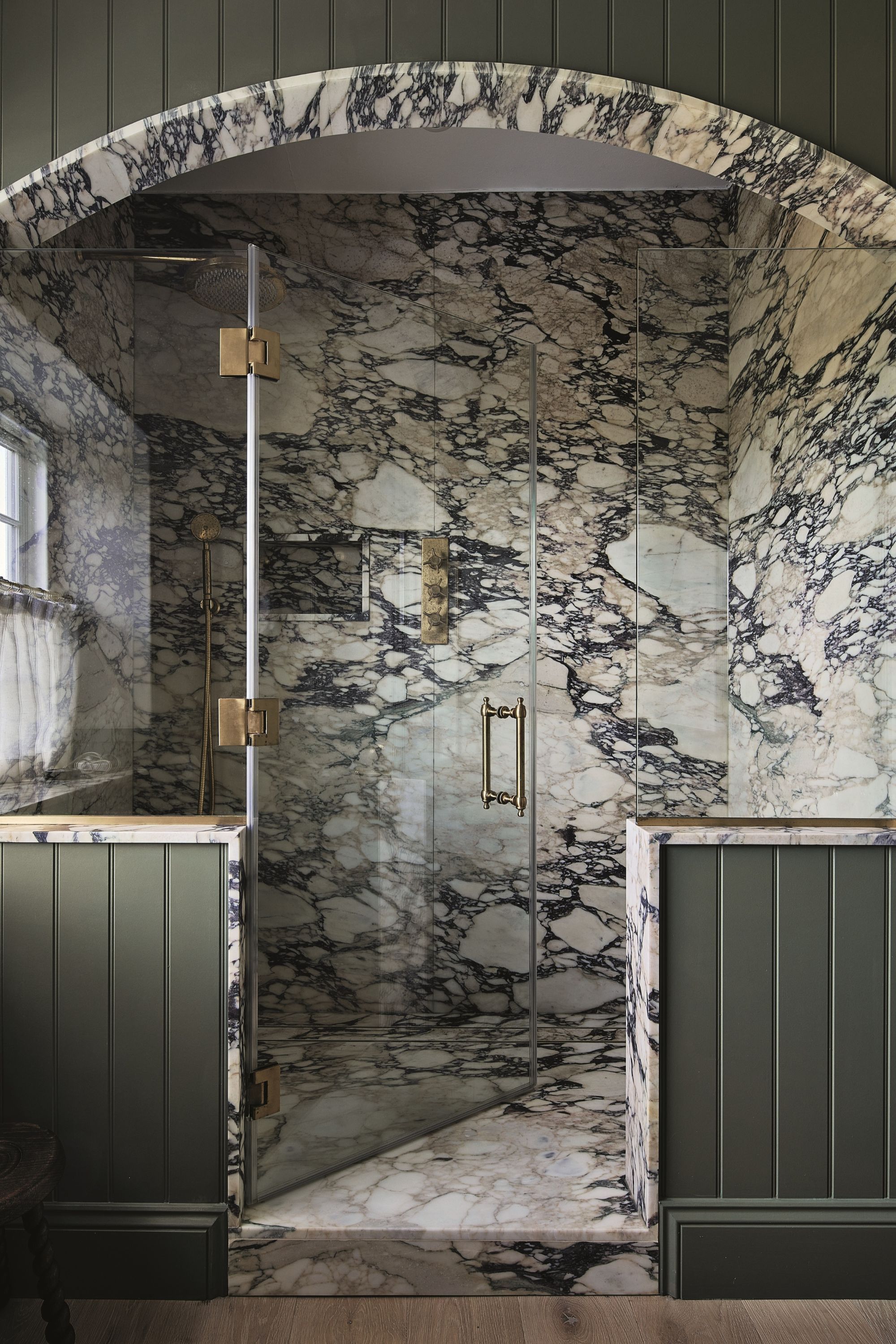 walk-in shower niche drenched in marble, with glass door and gold hardware, arch and green panelling with timber floorboards outside