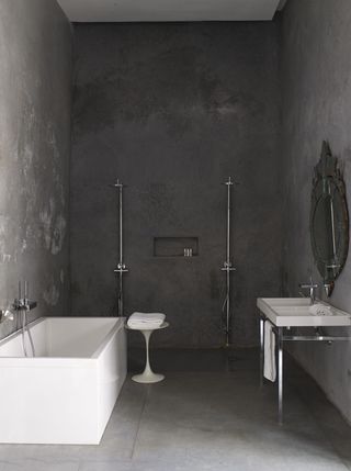 A hotel bathroom with a white tub, a white sink, a decorative mirror, twin silver showers, a round table and grey walls.