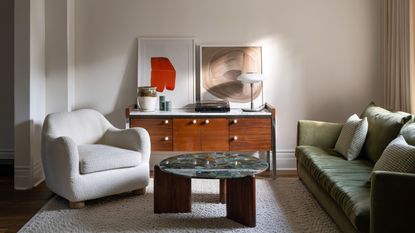 A warm, contemporary living room with an olive green sofa, sculptural cream armchair, wooden sideboard, statement art and a coffee table.