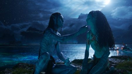 Still from the movie Avatar: Fire and Ash. Two blue, tall, slender Na'vi - Lo'ak (Britain Dalton) and Tsireya (Bailey Bass) - are sitting on the shore in the moonlight. Lo'ak is reaching out to hold Tsireya's chin.