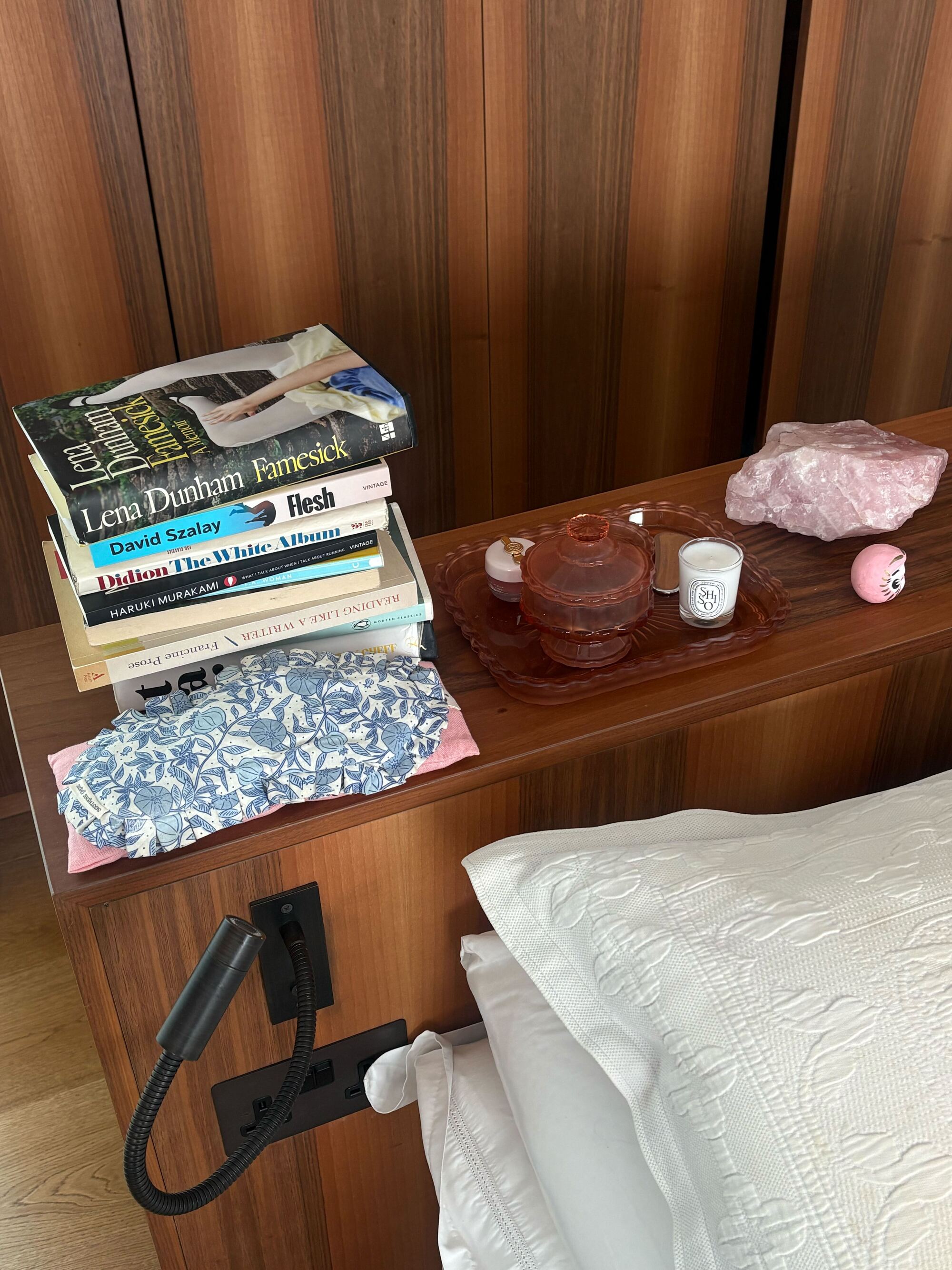 Image of a wooden bed with an extended headboard that has a stack of books, a blue and white eye mask, a pink decorative tray with a candle on it, and a pink rock.