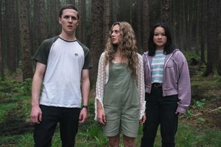 Jamie (Oscar Kennedy), Pippa (Jodie Tyack) and Vivian (Thaddea Graham) are standing in a clearing in the forest. Jamie and VIvian are staring straight ahead, Pippa is looking at Jamie with concern