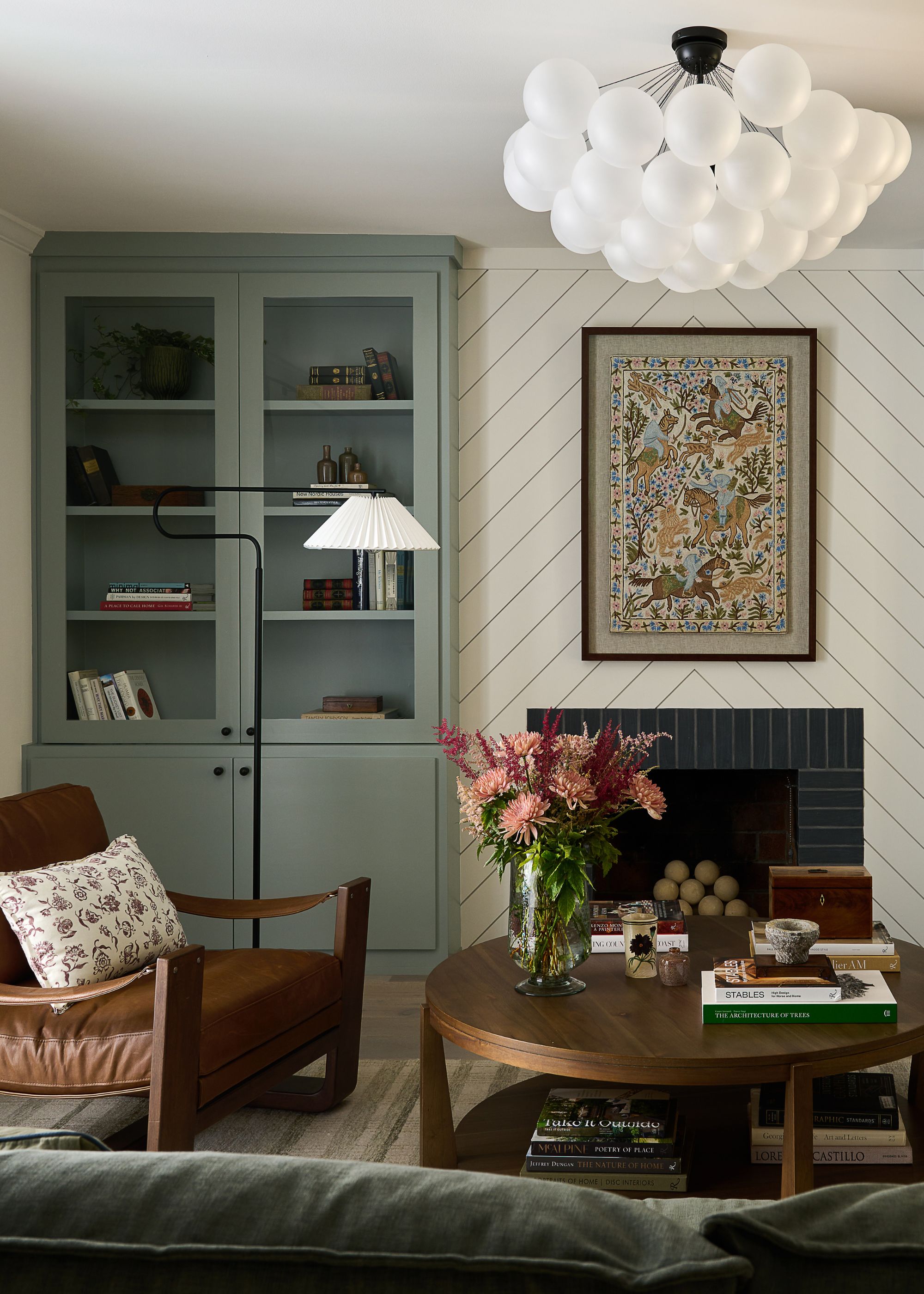 A cozy living room with white herringbone paneling above a fireplace, and a blue-green cabinet to the side. A round wooden coffee table and a leather accent chair.