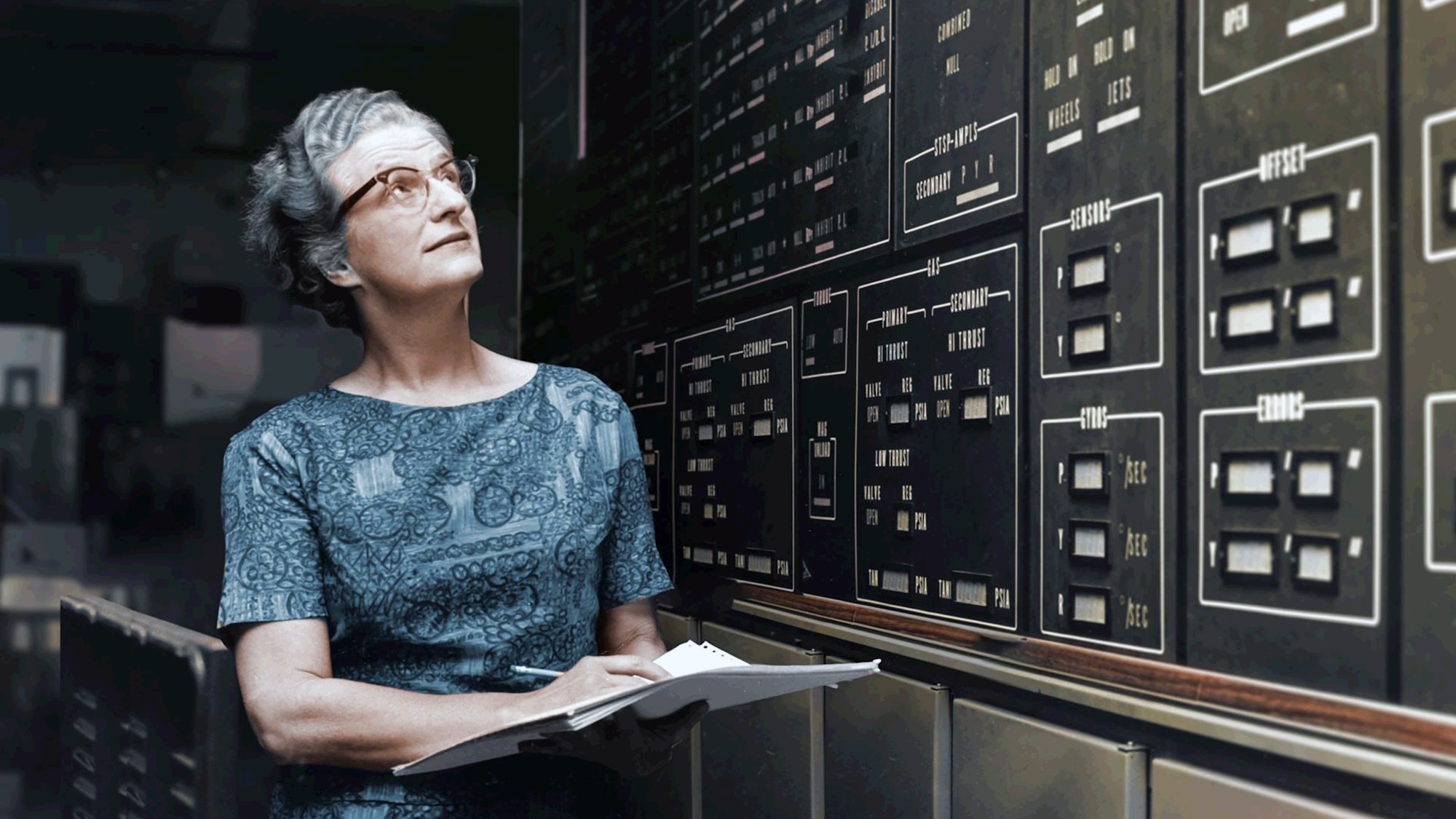 a woman in glasses carries a notebook and pencil beside a wall covered in instrument panels and lights