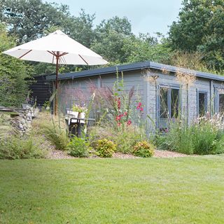 A Lillevilla garden cabin next to dining table set with cream-coloured parasol. Tall plants, such as Stipa gigantea and Verbena bonariensis planted throughout