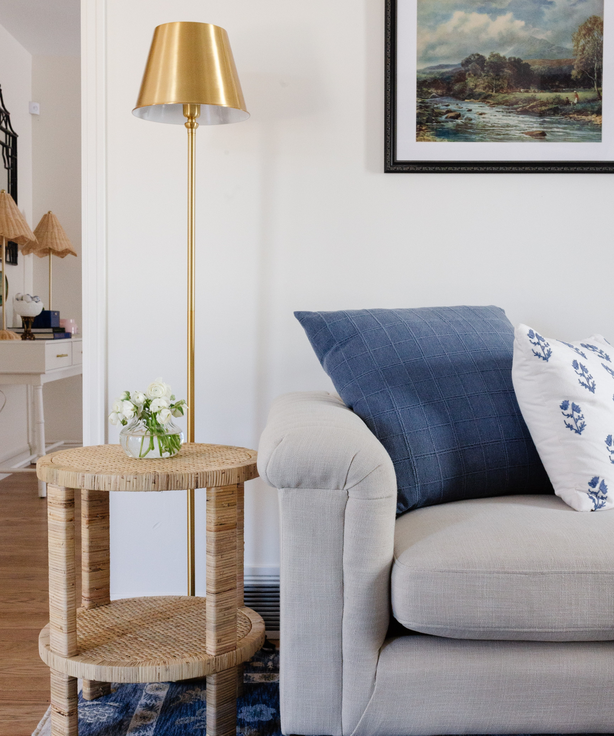 a living room with a grey couch with pillows, a small rattan side table, a gold floor lamp and a landscape image on the wall