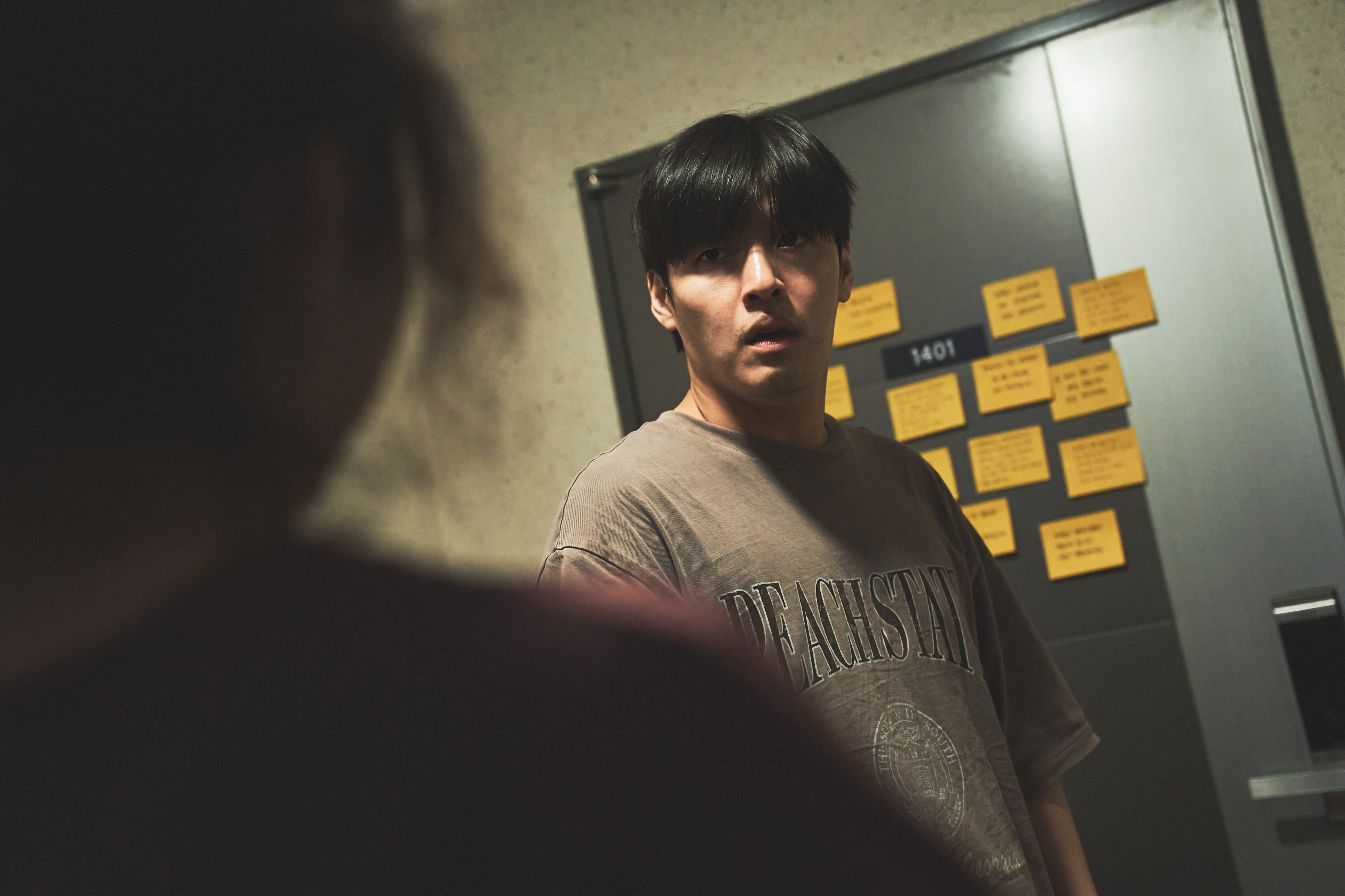 A man (Kang Ha-neul as No Woo-sung) looks in fear at an unseen woman as he stands in front of an apartment door covered in sticky notes, in the Netflix Korean movie 'Wall to Wall.'