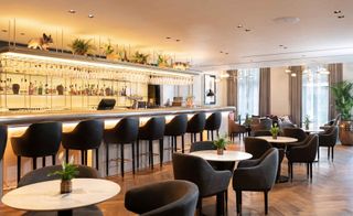 A bar inside the Hilton London Bankside hotel. To the left, there is a bar, which spans almost the entire room. We see shelves with drinks and glasses behind the bar. Bar chairs in black leather are next to the countertop. White marble tables with dark gray velvet chairs are set throughout the room. The entire place is very light and airy.