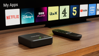 A Manhattan Aero 4K TV Streamer and remote on a wooden bench sitting below a TV screen which shows a list of streaming services.