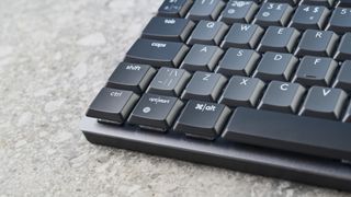 A photo of the Logitech MX Mechanical Mini keyboard in black and gray, on a stone slab and wooden table with a blue background.