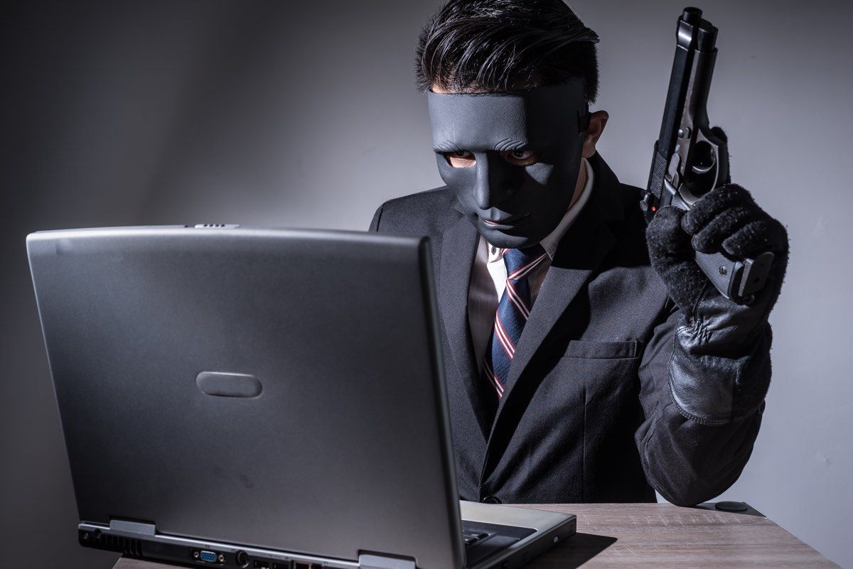 Abstract image of hacker or criminal wearing a mask and suit, using a laptop and holding a gun.