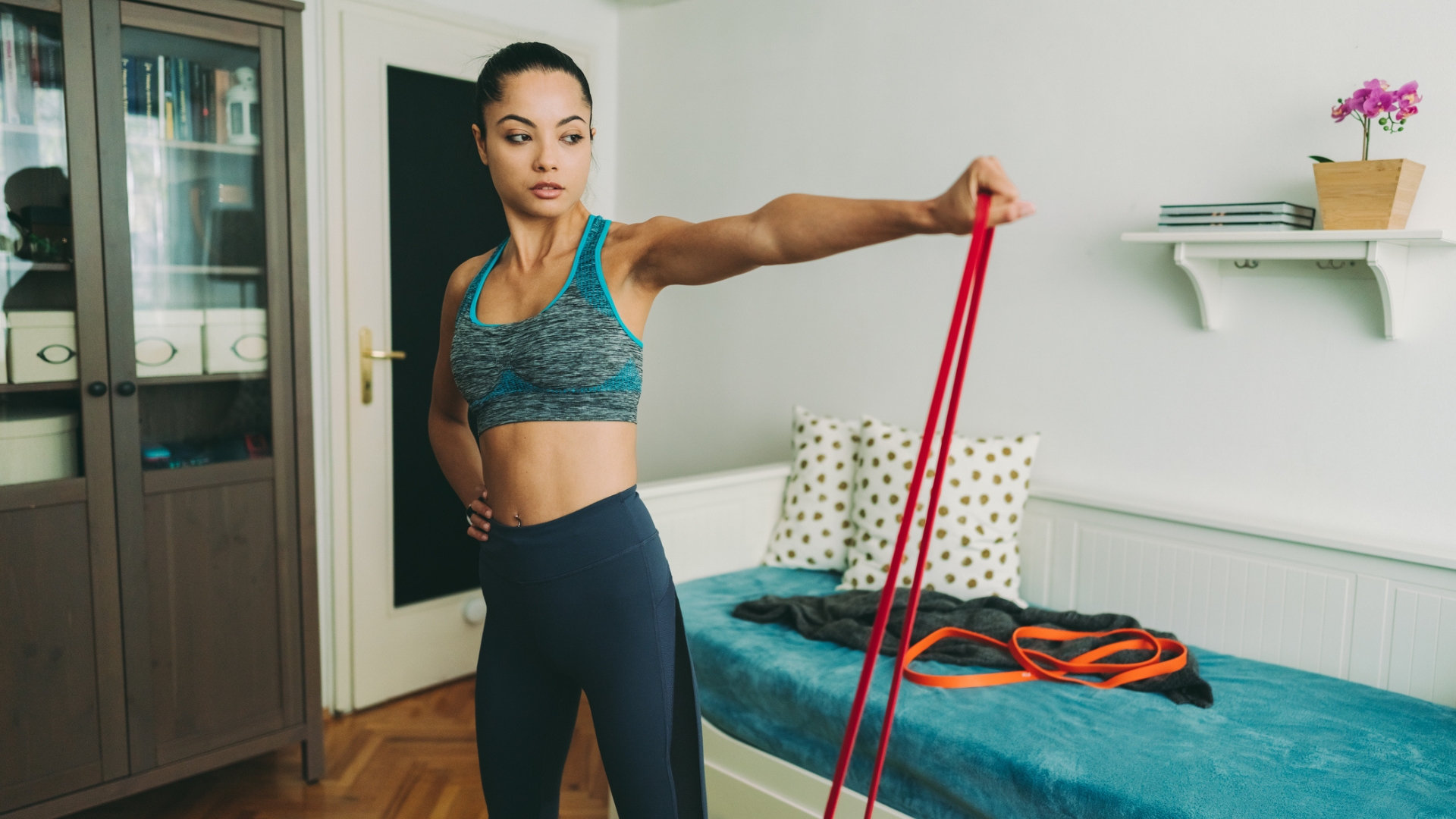 woman wearing a crop top and leggings holding a red resistance band to shoulder height and looking at it. she's in a home setting with a sofa behind her with other resistance bands 