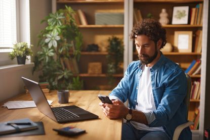 A man in his forties looks at his phone. A laptop is open on the desk in front of him. He is concentrating.