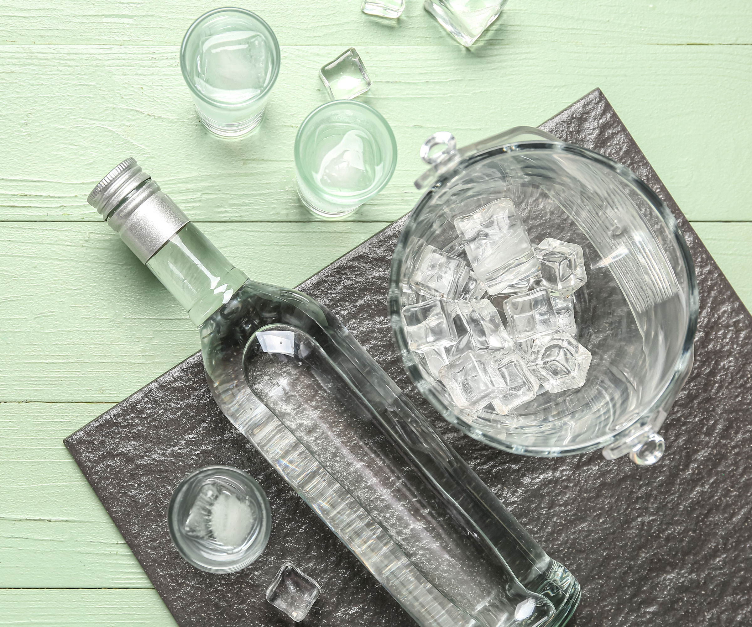 bottle of vodka and jug of ice with glasses on metal board with green wooden table