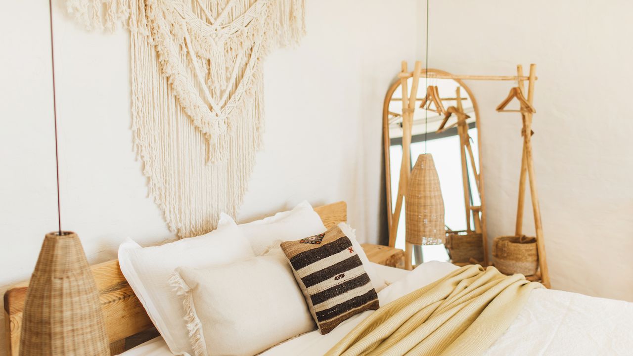 A closeup of a boho bedroom, particularly the bed, woven hanging light, a textile wall art piece, and a wooden clothing rack