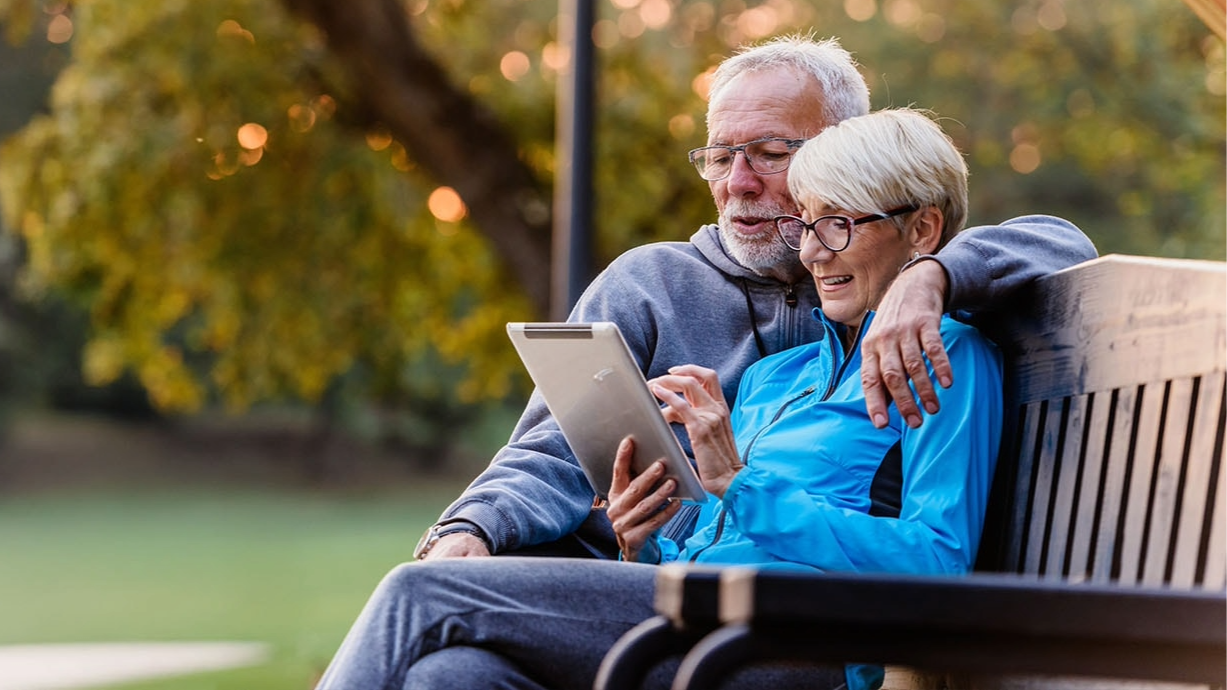 A couple sitting on a bench outside, looking at a tablet.