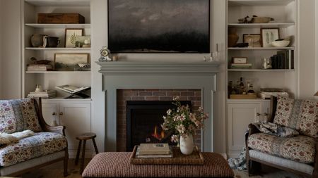 Cozy living room with soft white walls and built-ins with collected decor on show. A stone-colored fireplace with a landscape artwork above it. Two armchairs with a ditsy floral pattern and an ottoman