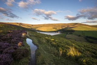 A puddle on a footpath leading along a hillside leading to Kinder Scout with Kinder reservoir below.