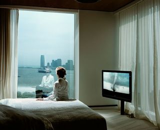A woman sitting on a bed looking out of a window. There is a television with a tornado on the screen