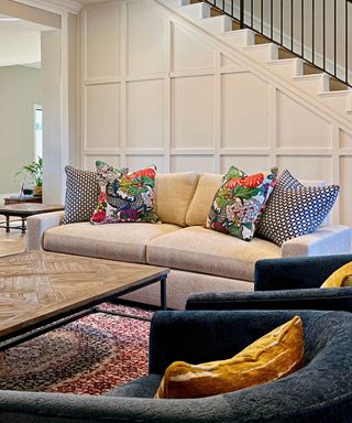 A living area with cream wall panels on the stairs, a cream couch with five throw pillows in navy blue, yellow, and multicolor, a dark wooden rectangular coffee table, and two curved navy blue arm chairs with yellow throw pillows on them and a multicolored rug on the floor