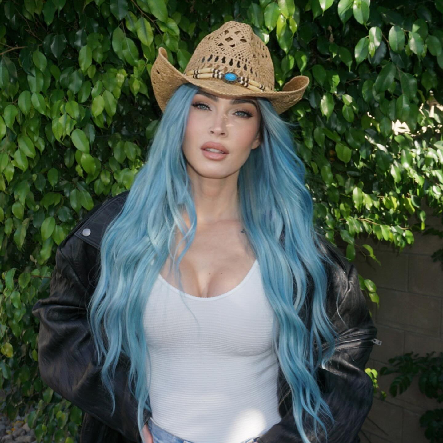 Megan Fox posing for a picture in front of greenery at Coachella wearing a cowboy hat, white tank, and black jacket