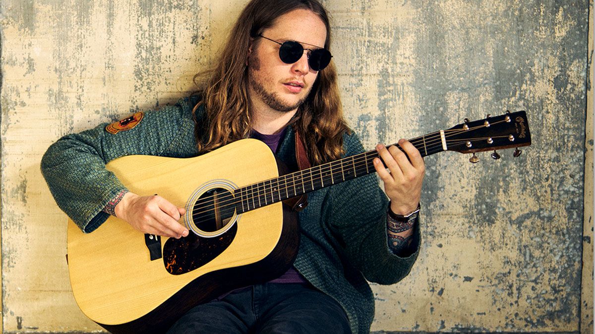 Billy Strings wears dark sunglasses as he sits against a wall and plays his new signature Martin D-28 acoustic guitar