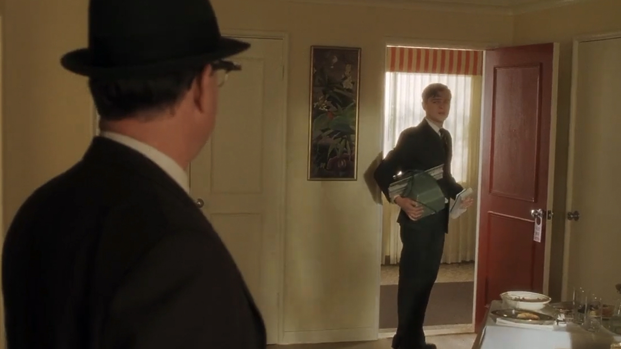 Tom Hanks in a fedora with his back to the camera looking Leonardo DiCaprio standing in a doorway with a machine in his hand.