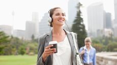 Woman smiles as she walks with a coffee in hand listening to music through headphones