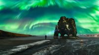 A person stands on a dark beach under a vibrant display of green and blue northern lights, which streak across the sky in a dynamic, swirling pattern. A large, rugged rock formation rises nearby, adding to the dramatic scene.