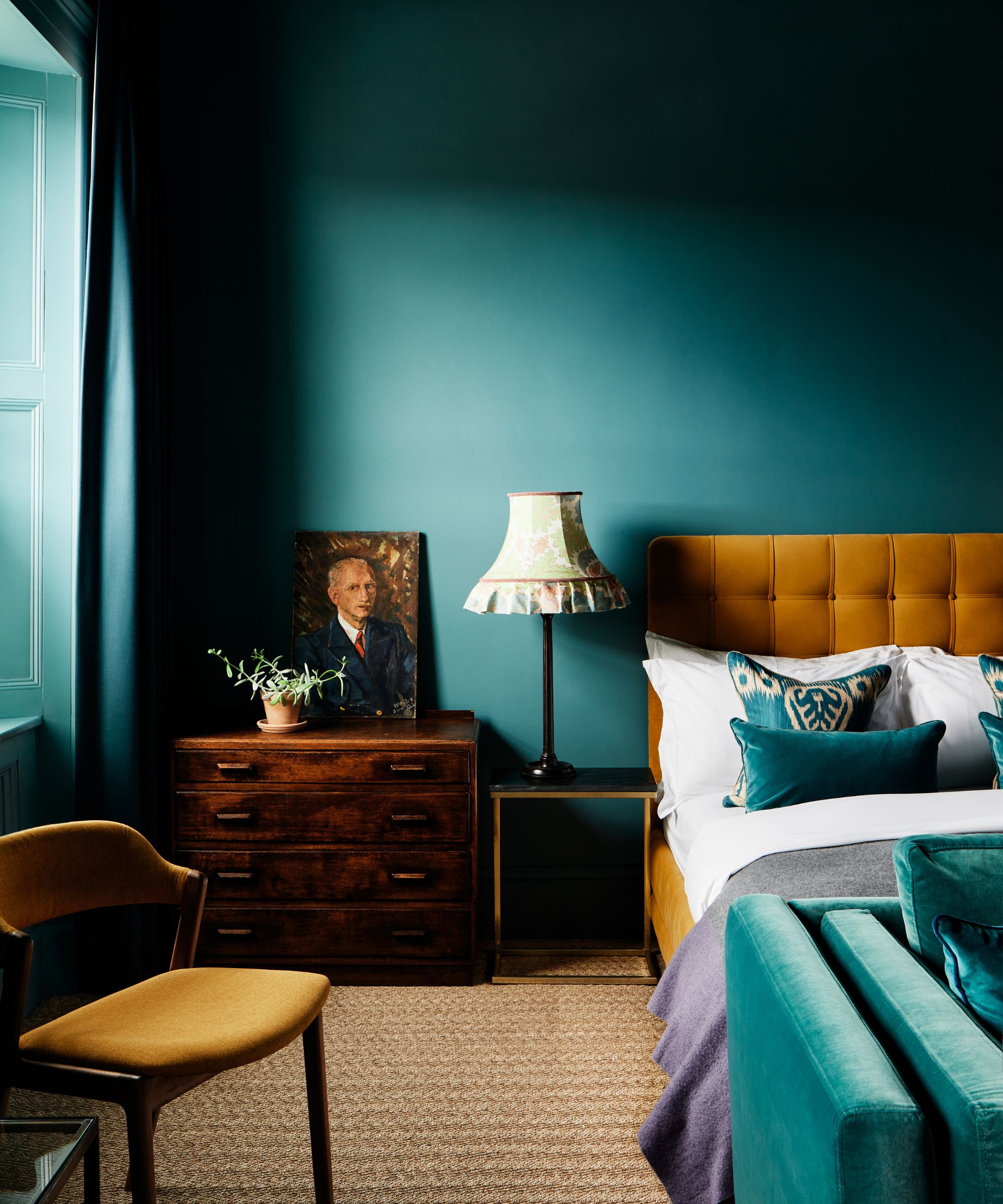 A bedroom with color-drenched teal walls, a mustard bed with white and teal bedding, and a wooden chest of drawers with artwork on it.