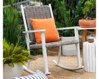 A wicker and white aluminum outdoor rocking chair with an orange pillow in the garden
