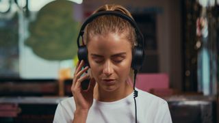 A woman in a white top with her blonde hair scraped back is listening to something on a pair of RØDE NTH-50 headphones with her eyes closed. 