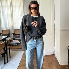 French fashion influencer Anne-Laure Mais poses for a mirror selfie in her chic Paris apartment with chevron wood floors wearing black sunglasses, a charcoal gray crewneck sweater layered over a white T-shirt, a mini Louis Vuitton chain shoulder bag, a black belt, and light-wash jeans 