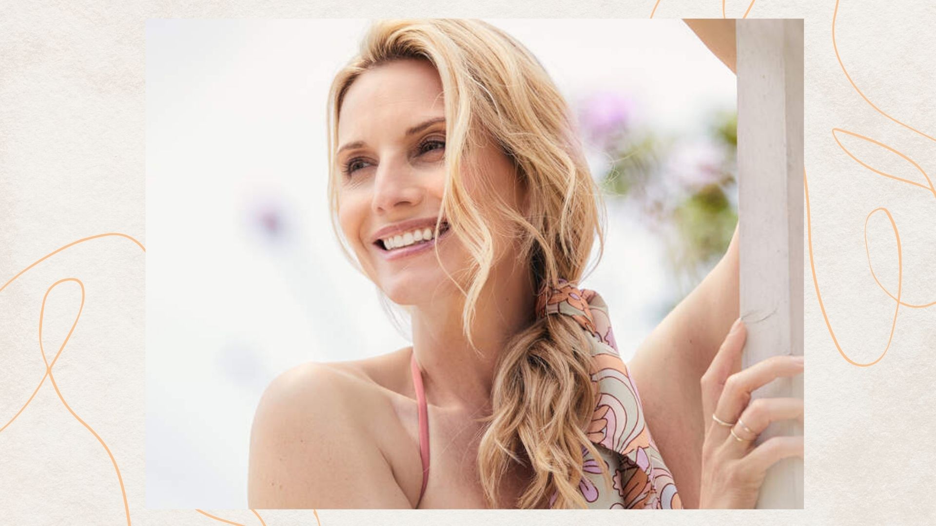 a woman with blonde wavy hair with a scarf tied around it, pictured outdoors in the sunshine with flowers in the background