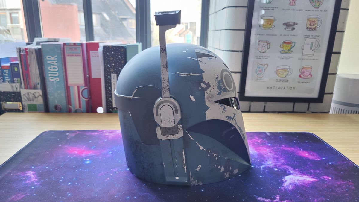 The Star Wars: Black Series Bo-Katan Helmet with the heads-up display up. There is a gray, black, and white owl-like pattern on the front of the light blue helmet and a black T-shaped visor. This photo shows the right-side of the helmet.