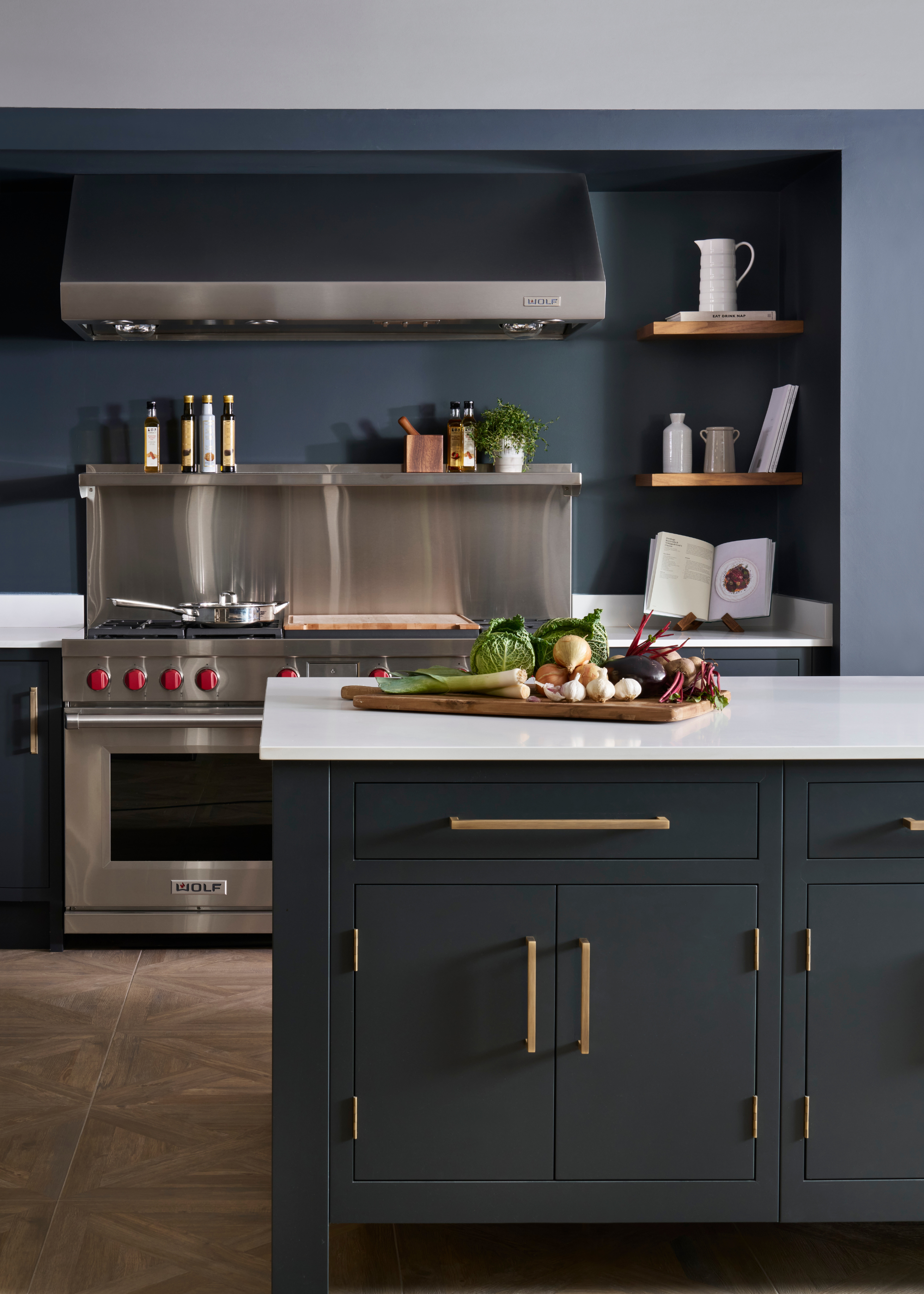 A dark blue kitchen with large stainless steel cooker, splashback and extractor, open shelving and fresh veg on a wood board on the island